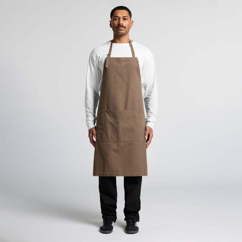 A man stands wearing a brown canvas apron over a white shirt, featuring a logo and front pockets.