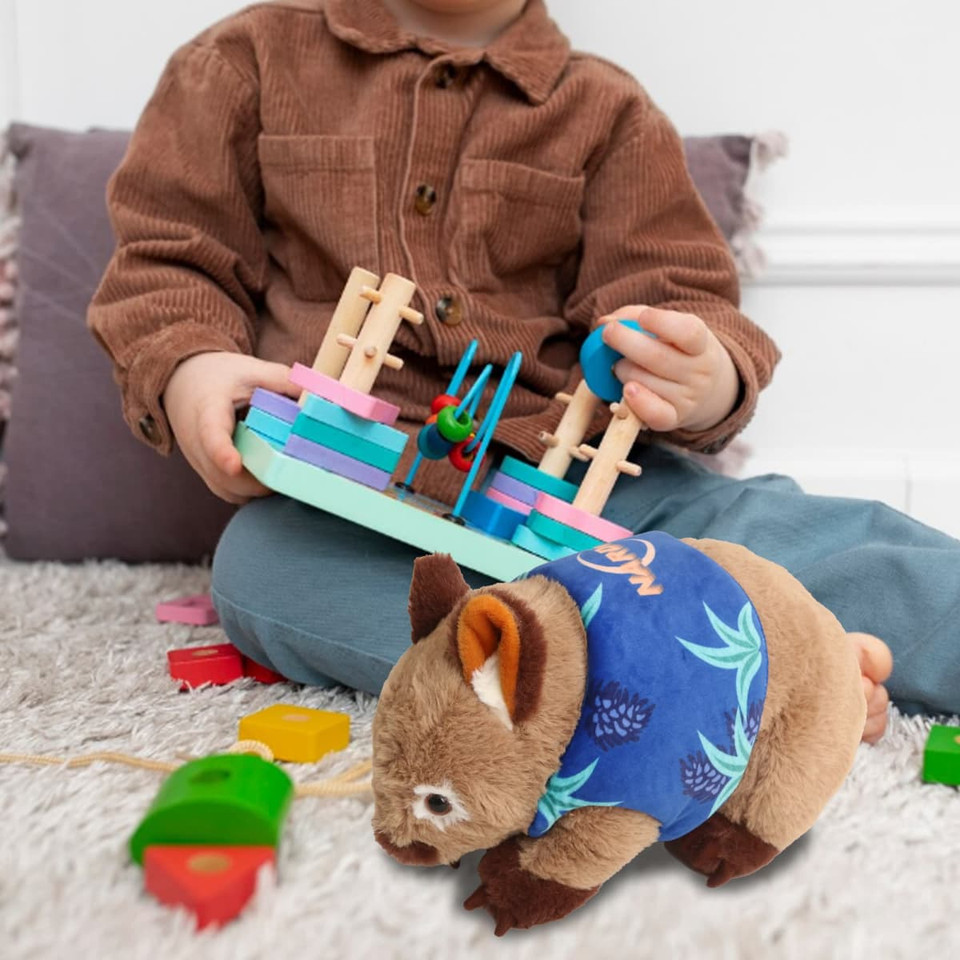 A plush wombat toy with a blue shirt sits on a soft surface, beside a child playing with colourful blocks.