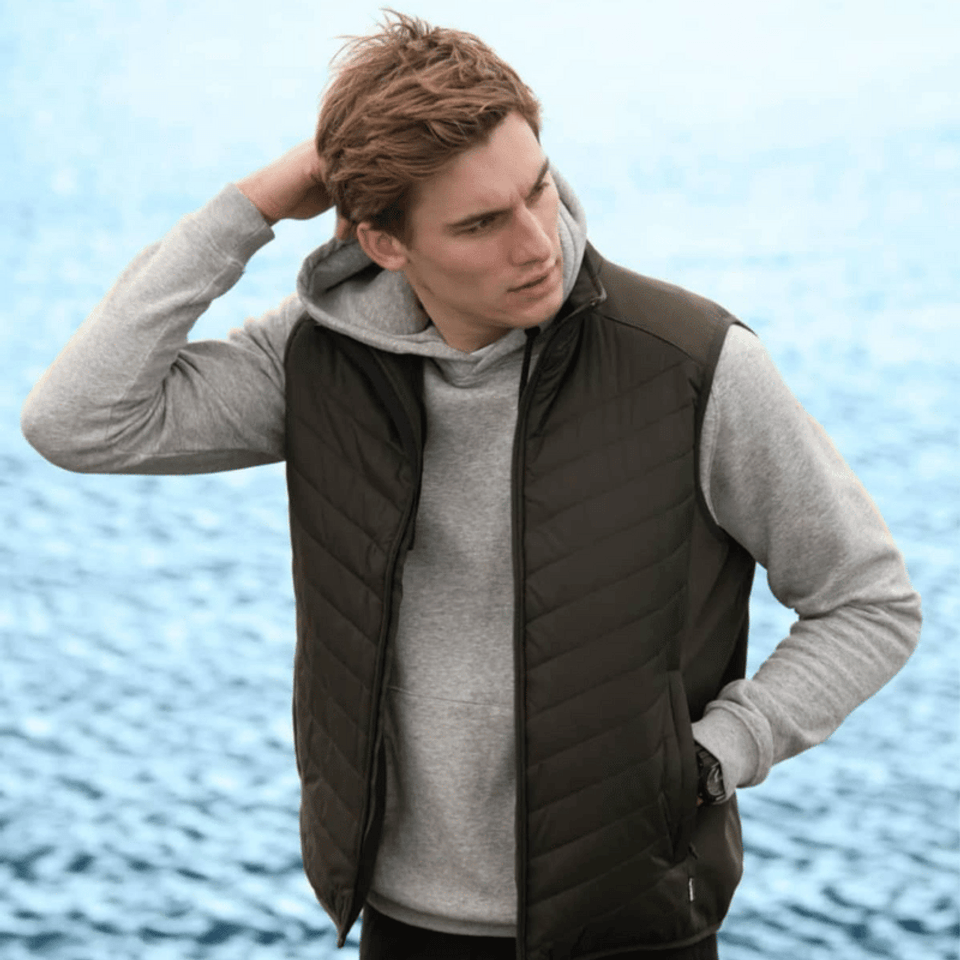 A green puffer vest worn over a grey hoodie, modelled by a young man against a water background.