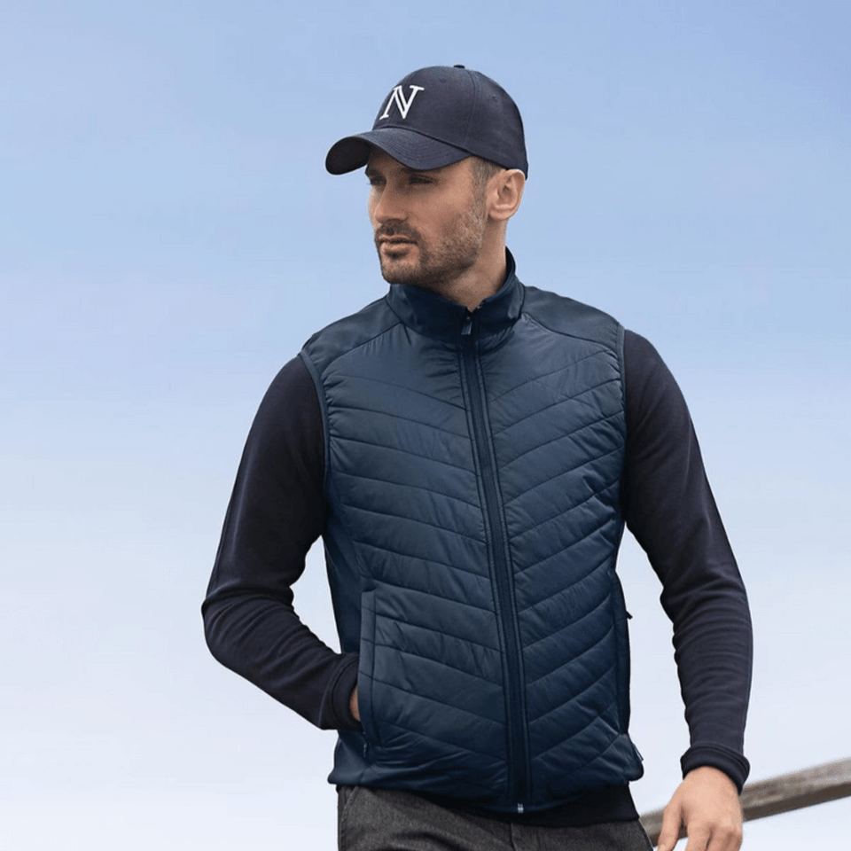 A navy blue men's vest with a quilted design, featuring a zip and side pockets, worn with a black long-sleeve top and cap.