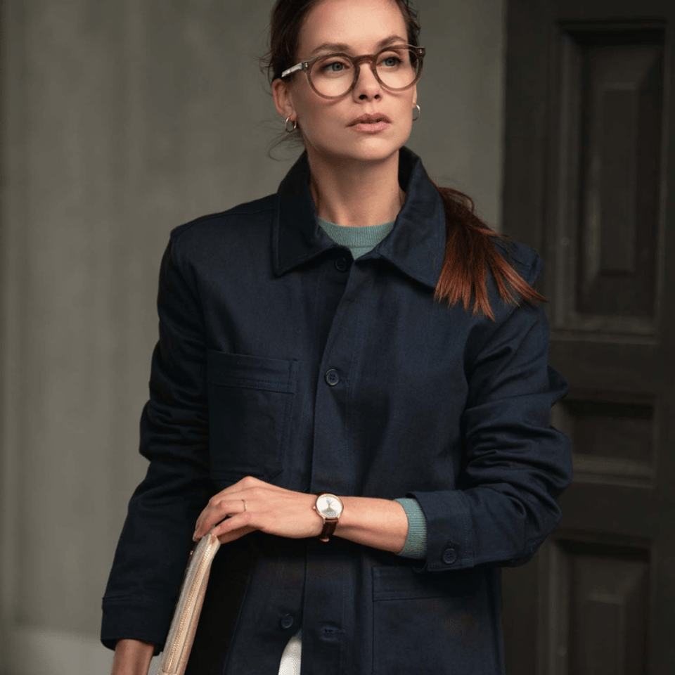 A woman wearing a dark navy jacket with a green top and white pants, holding a notebook and walking.