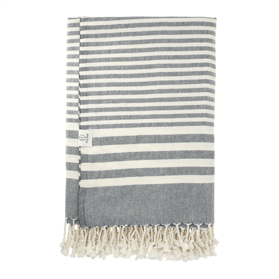 A grey and white striped blanket with tassel detailing, featuring a small logo tag.