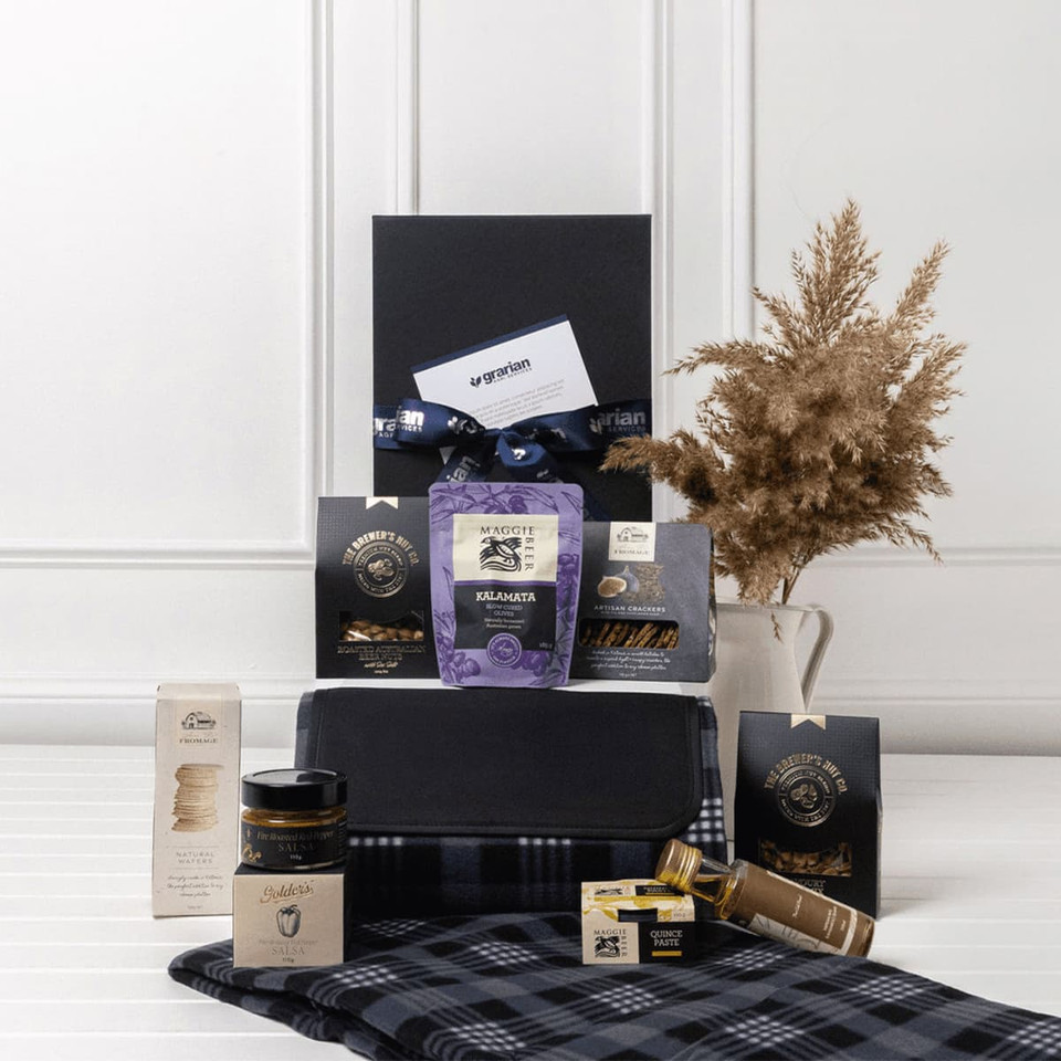 A stylish black gift box containing gourmet snacks, a picnic blanket, and packaged treats, featuring a decorative arrangement.