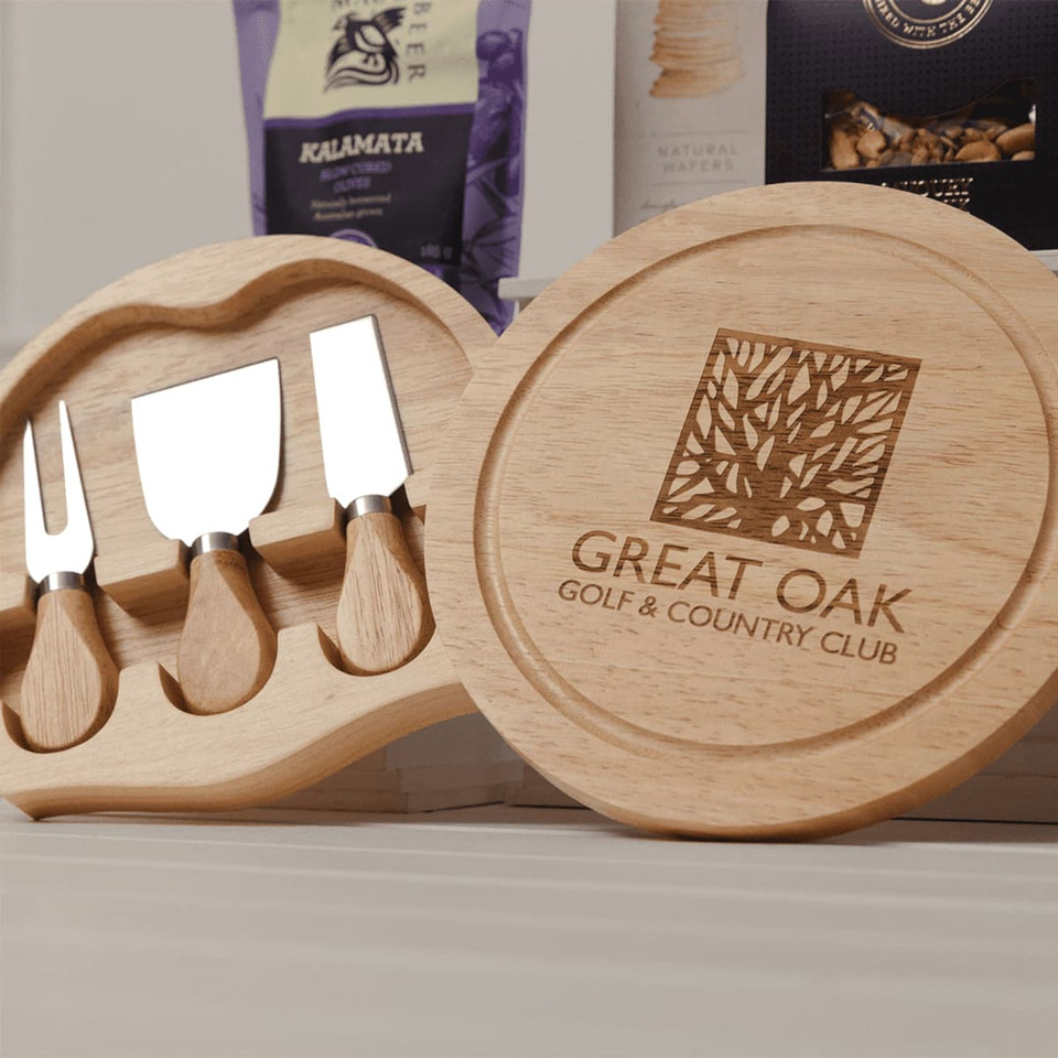 A wooden platter containing three cheese knives, featuring a logo, alongside gourmet snacks.
