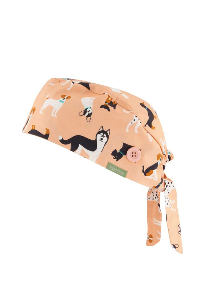 A unisex scrub cap in a light peach colour, featuring a pattern of various dogs and a button detail.