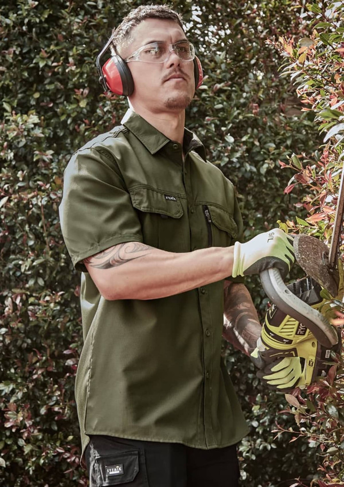A man wearing an olive green short sleeve button-up shirt, safety glasses, and ear protection, using a power tool.