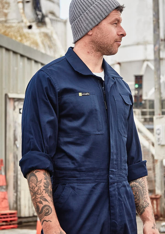 A man wearing a navy blue lightweight cotton drill overall with a logo on the chest, standing in an industrial setting.