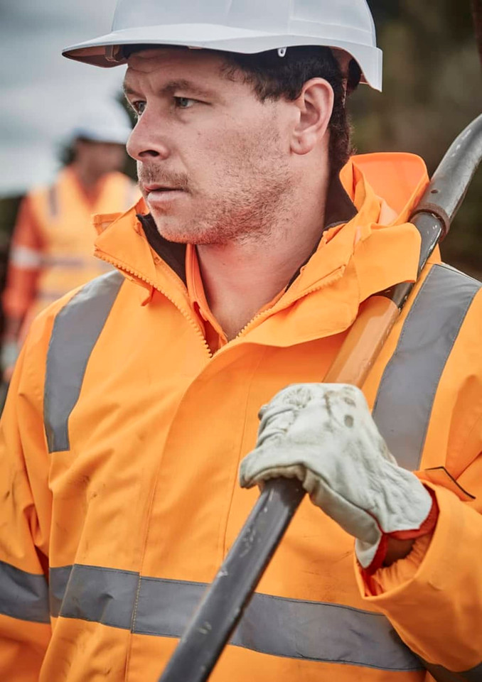 A man wearing an orange waterproof jacket with reflective strips, gloves, and a safety helmet, holding a tool.