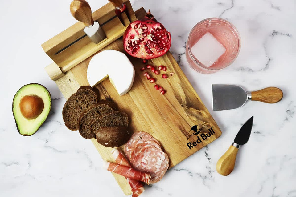 A wooden cheeseboard with cheese, meats, avocado, bread, and a pomegranate, featuring utensils and a logo.