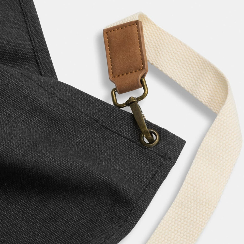 A black canvas apron featuring a leather strap and a secure metal clip. It has a cream-coloured shoulder strap.