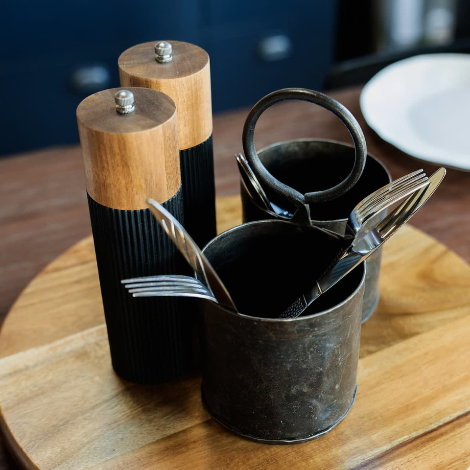 Keepsake Noir salt and pepper grinders in black and wood, alongside utensils in a metal holder on a wooden tray.