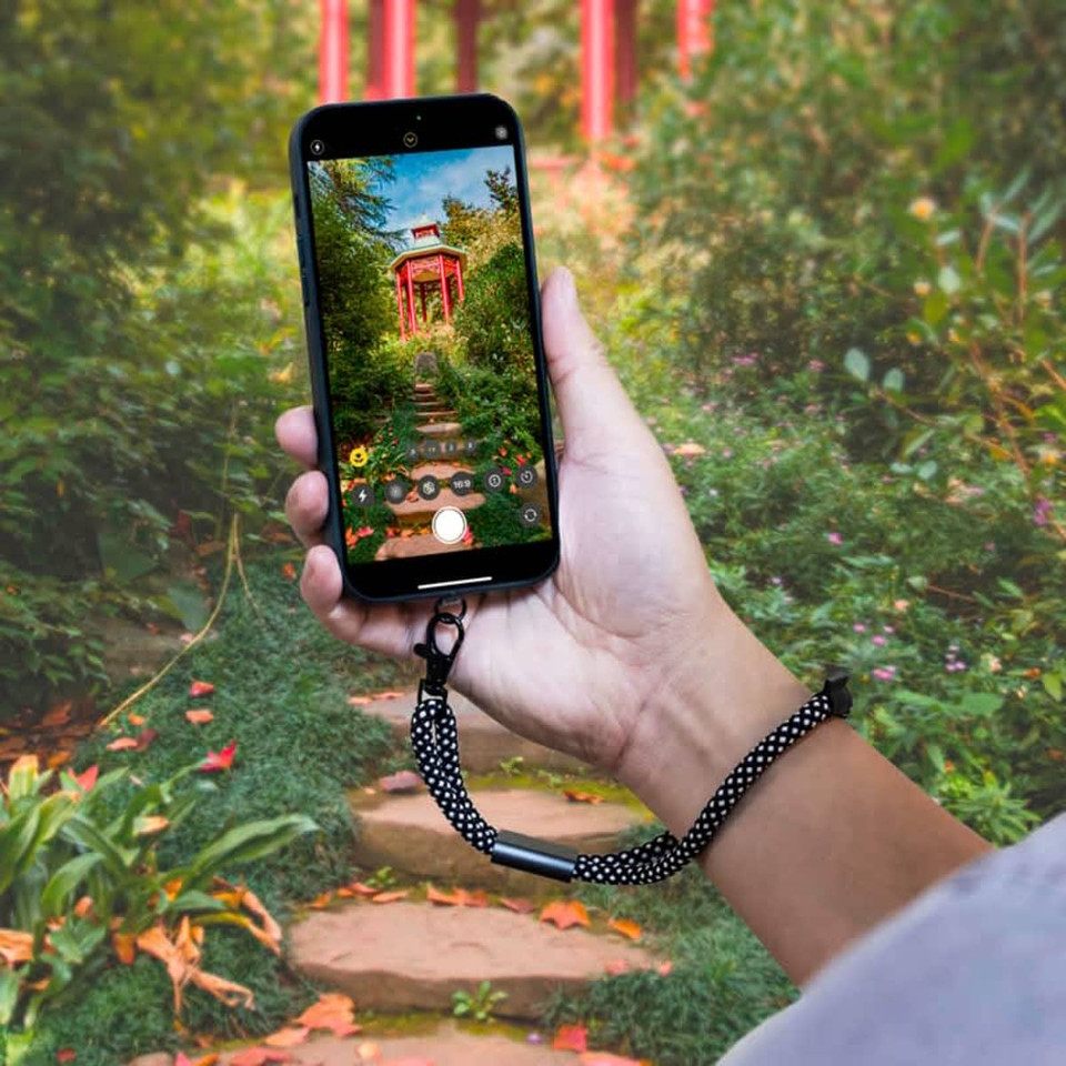 A smartphone is held by a hand with a black and white polka dot wrist strap, set against a lush garden background.