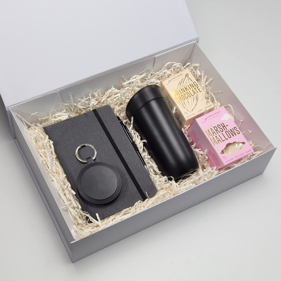 A travel mug in black, a notebook, a keyring, and marshmallows in a gift box with shredded fill.