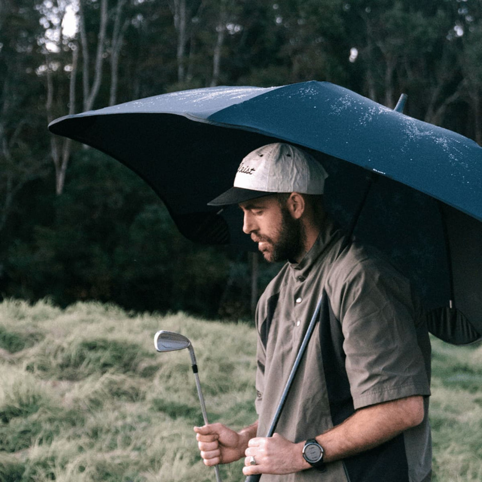 A black sport umbrella held by a man wearing a cap and grey shirt, standing on grass with trees in the background.