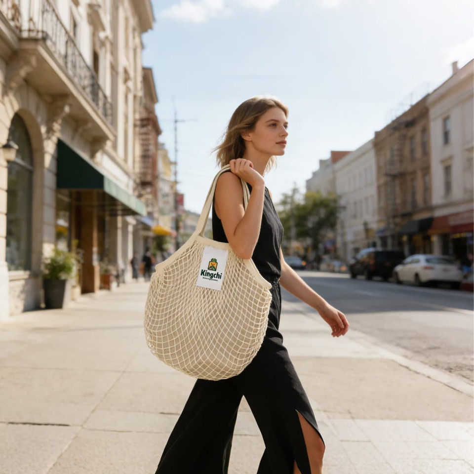 A woman walks along a street carrying a cream mesh grocery bag, featuring an inner section and a logo.