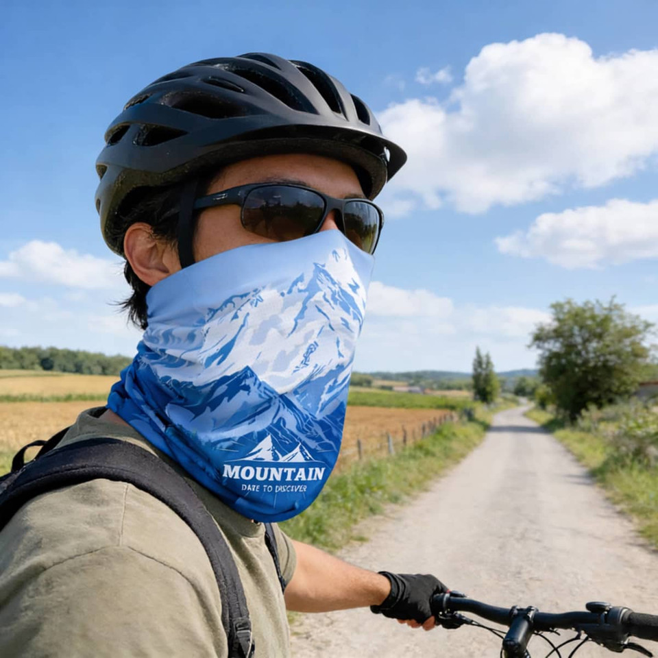 A cyclist wearing a blue UPF 50+ perforated ice silk neck gaiter featuring mountain designs against a rural backdrop.