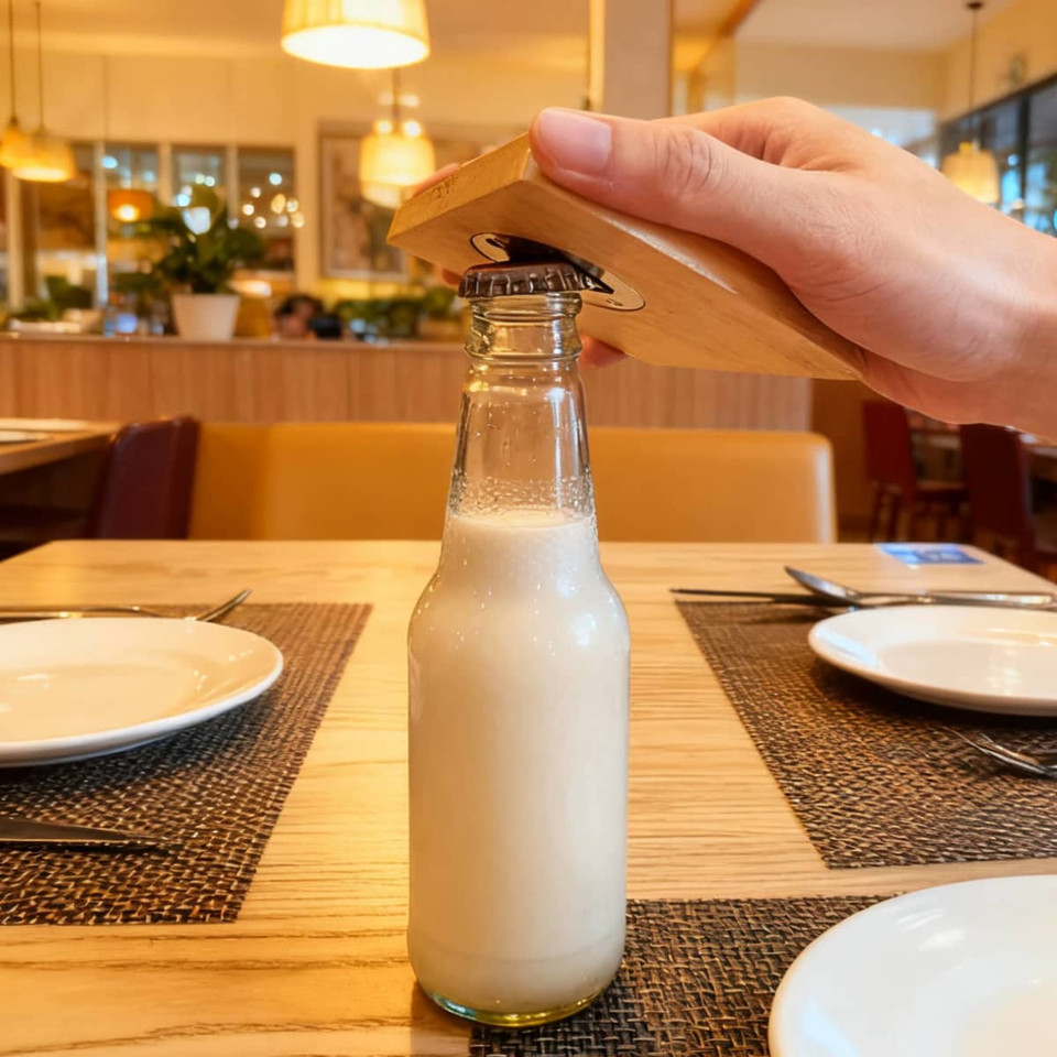 A drink bottle is being opened with a square bamboo bottle opener coaster on a wooden table in a restaurant setting.