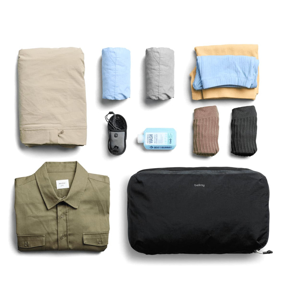 A black packing cube contains folded clothes, including shirts, pants, and socks, alongside a small bottle and a cable.