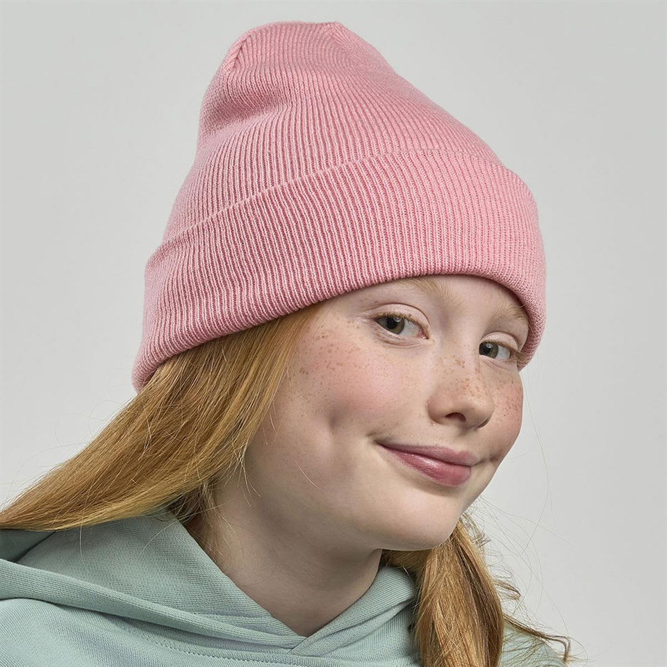 A pink ribbed beanie worn by a girl with red hair and freckles, featuring a logo on the side.