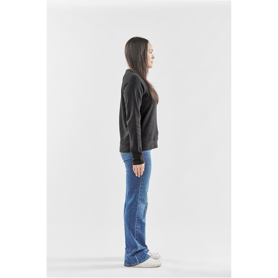 A woman stands sideways, wearing a dark grey crew neck fleece jumper and blue flared jeans, with white sneakers.