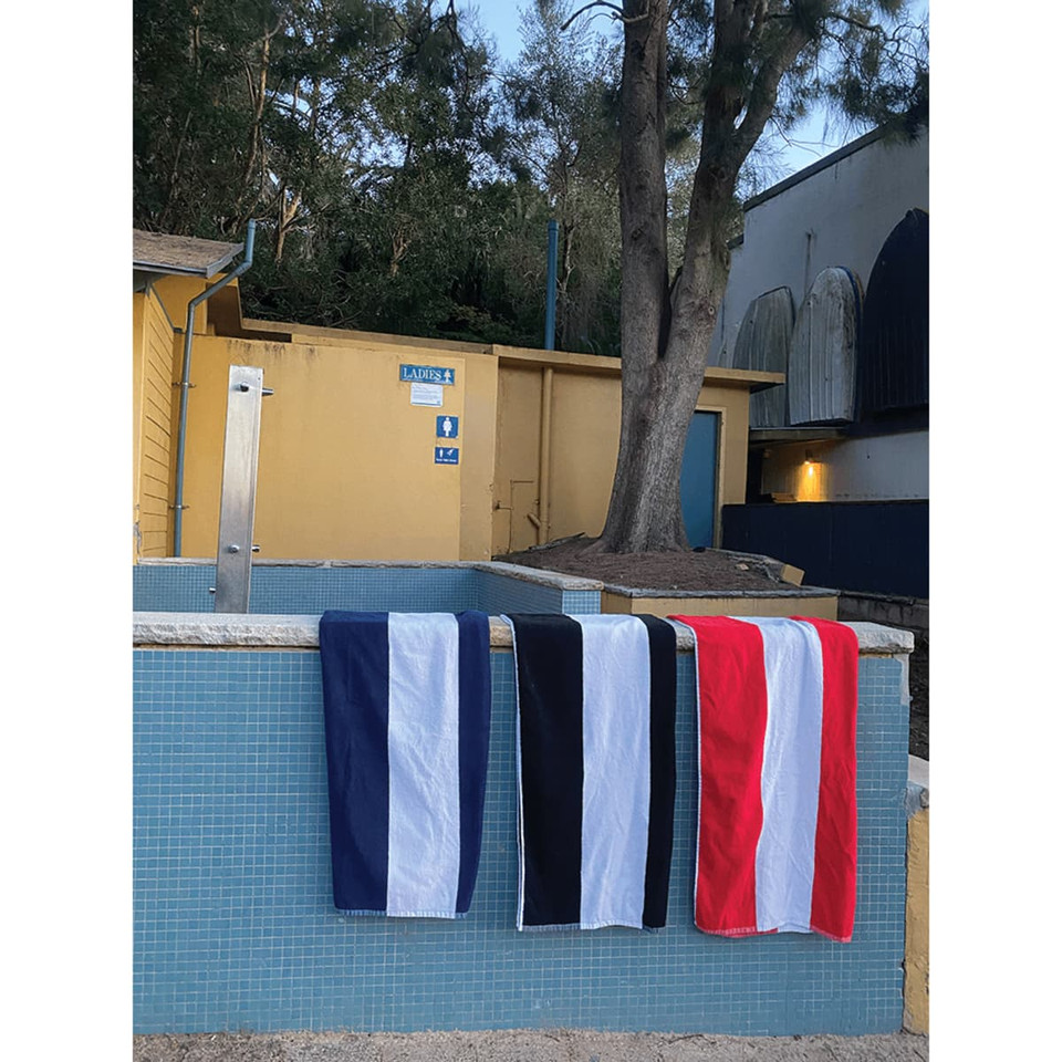 Three cotton beach towels in navy, black, and red hang on a railing near a pool, surrounded by greenery.