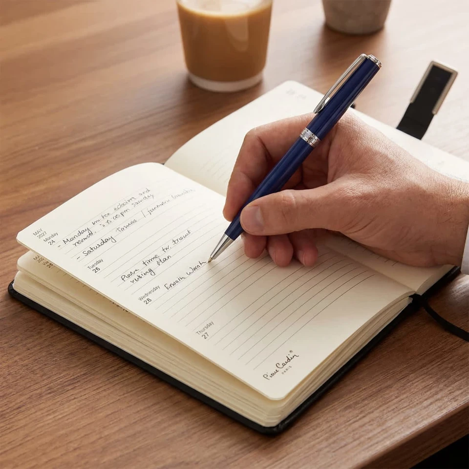A diary with cream-coloured pages and a pen resting on a wooden table, alongside a coffee cup.