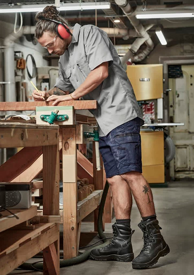 A man wearing grey work shirt and navy shorts stands at a workbench, using a pencil to mark wood. He has ear protection on.