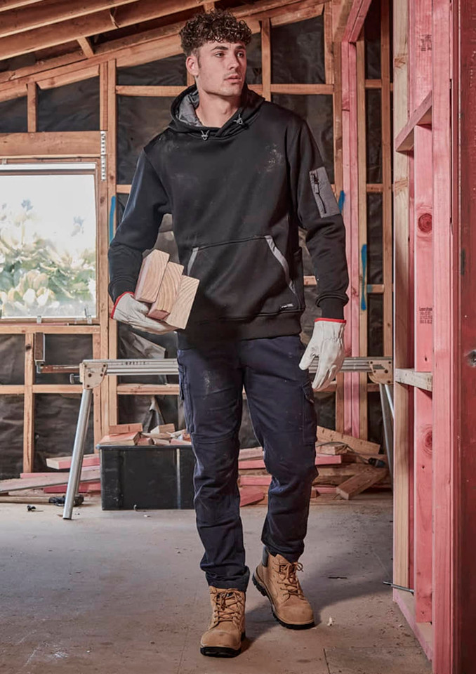 A man wearing black cargo pants and a black hoodie is holding wooden blocks, standing in a construction setting.