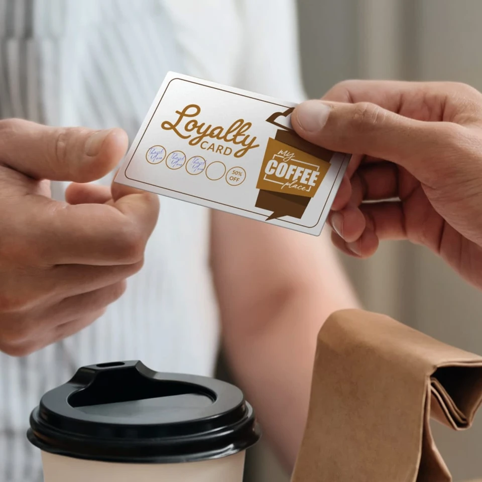 A hand holds a silver and gold loyalty card, next to a coffee cup and brown paper bag. A logo is visible on the card.