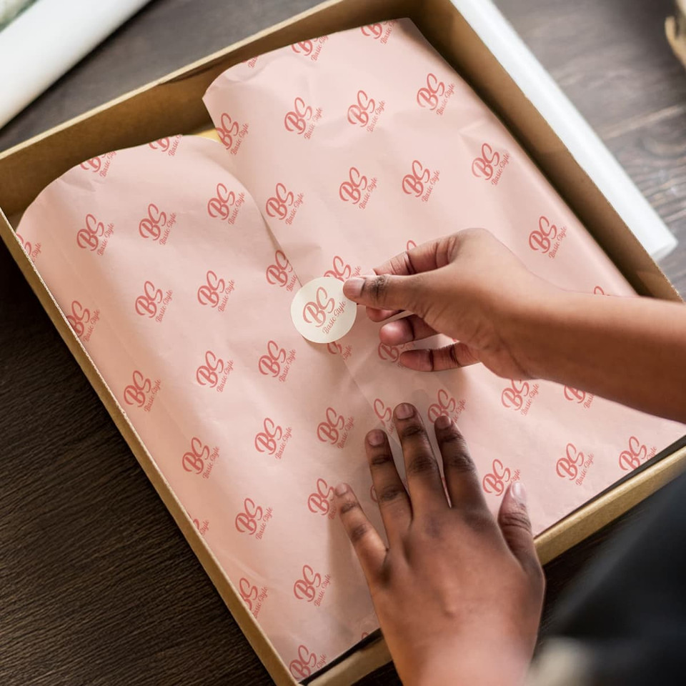 Tissue paper in pale pink with repeated branding, neatly placed inside a brown box. A hand is applying a sticker on top.