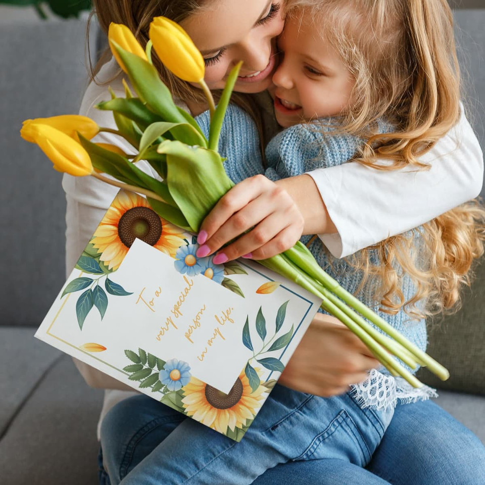 A woman hugs a girl while holding yellow tulips and an A4 message card featuring floral designs.