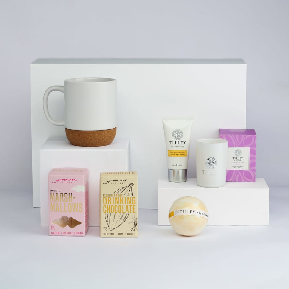 A white mug with a cork base, marshmallows, drinking chocolate, a candle, body cream, and a bath bomb in various packaging.