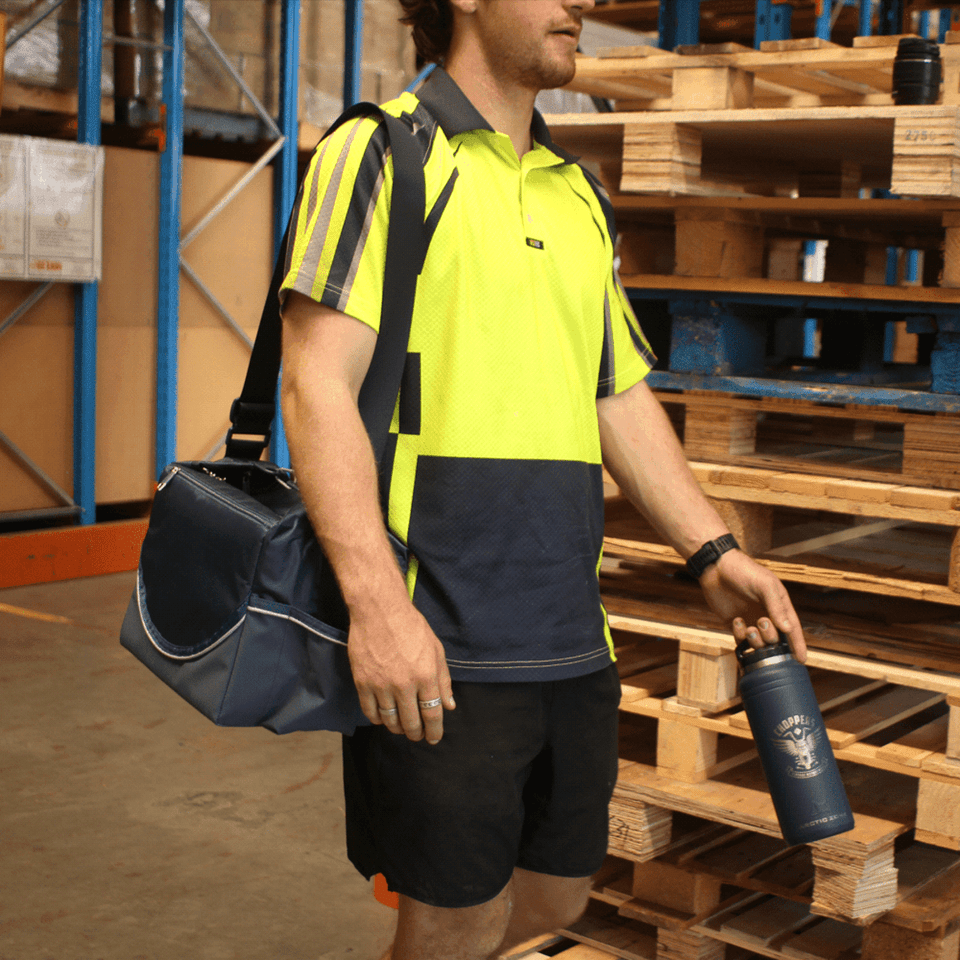 A man holds a navy cooler bag and drink bottle, wearing a bright yellow and navy sports shirt.