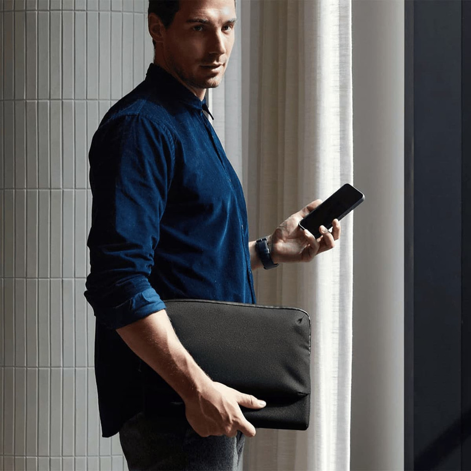A man in a navy shirt holds a smartphone and a black laptop caddy, standing by a window. The product has a logo.