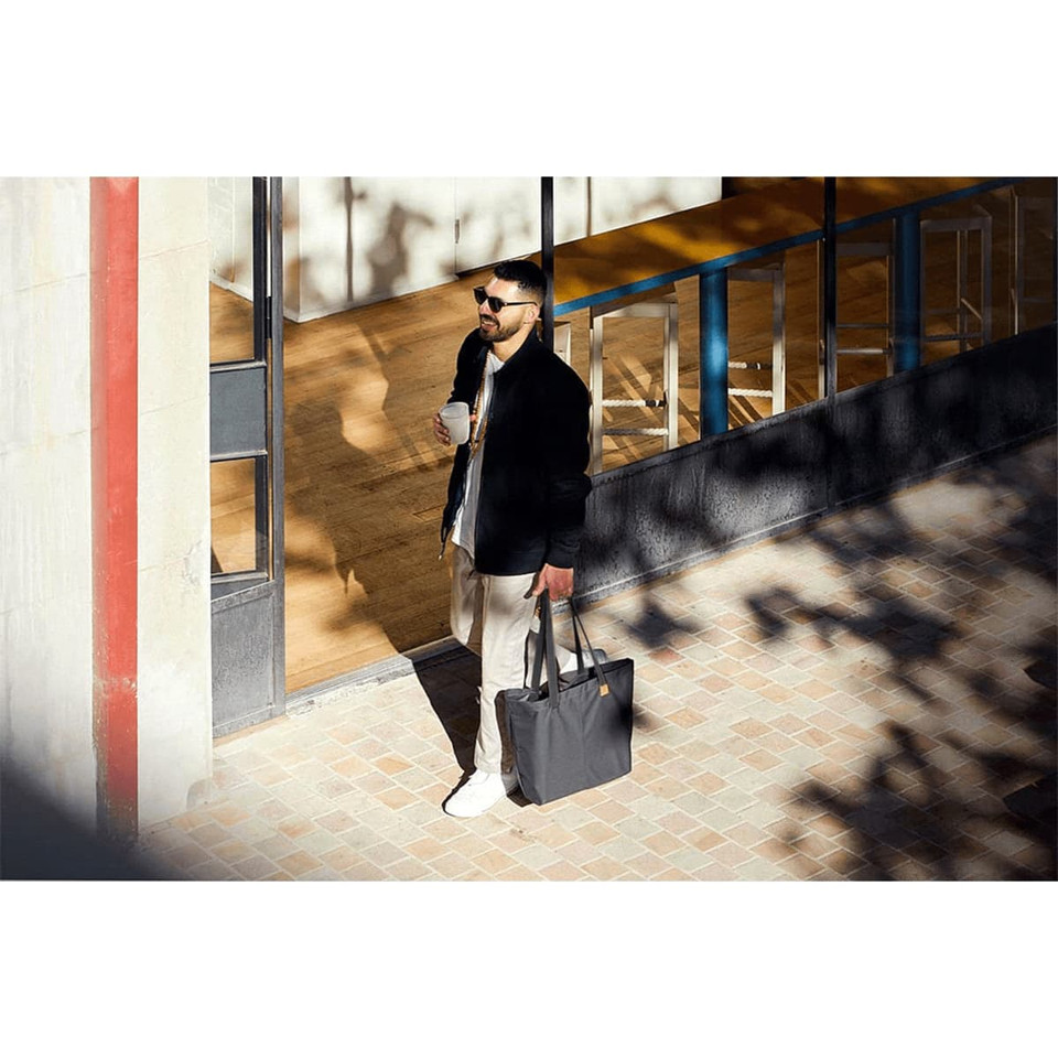 A man carries a dark grey tote bag while walking outside a building, wearing sunglasses and holding a drink.
