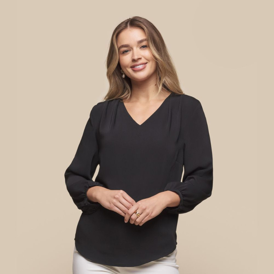 A female model wearing a black V Neck poppover shirt with three-quarter bell sleeves and pleated details, featuring a logo tag.