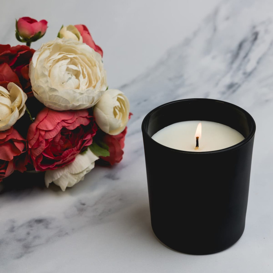 A large black glass candle with a white wax interior, placed beside a bouquet of red and cream roses.