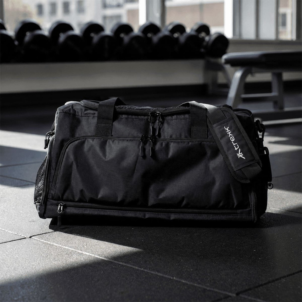 A black duffle bag with multiple zippered compartments and a logo, resting on a gym floor.