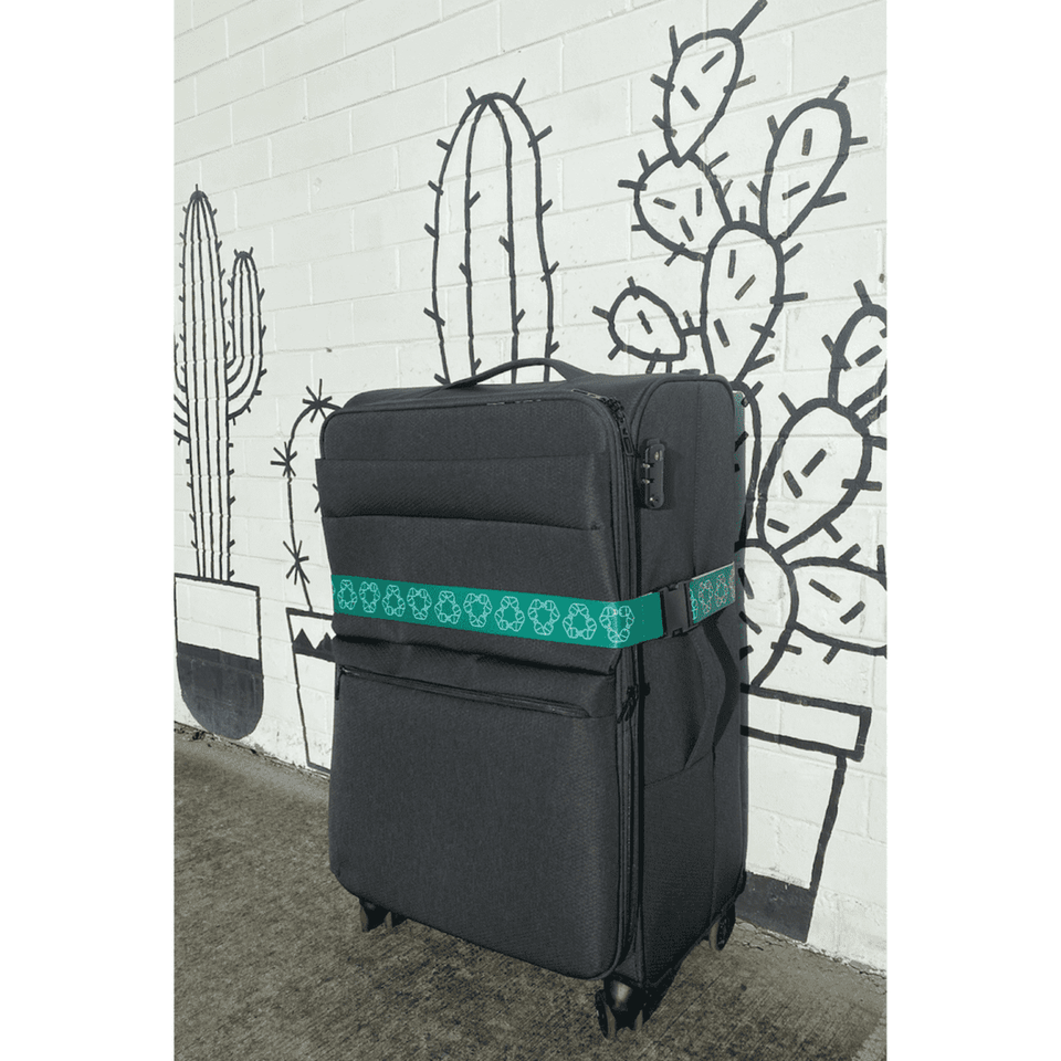 A black suitcase secured with a green rPET luggage strap, set against a wall featuring cactus illustrations.