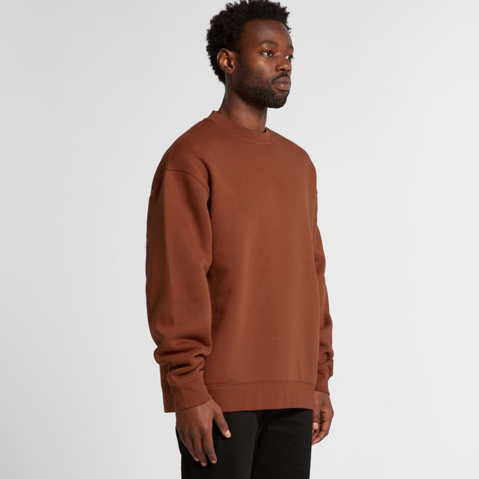A man wearing a brown sweater stands sideways, showcasing the relaxed fit and style of the garment.