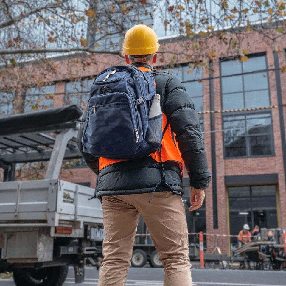 A person wearing a yellow hard hat and an orange vest carries a navy blue High Sierra Access 3.0 Backpack.