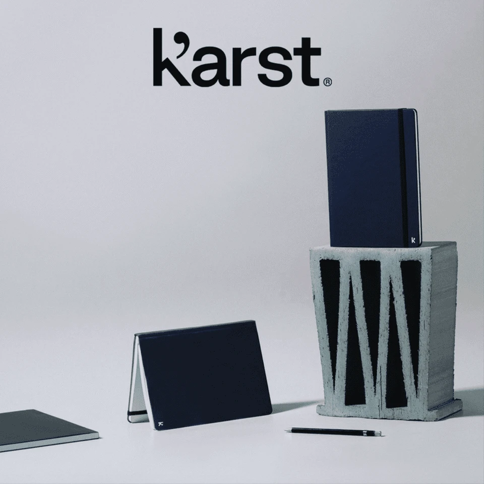 A navy hardcover notebook stands on top of a grey block, accompanied by a smaller notebook and a pen. It has a logo.