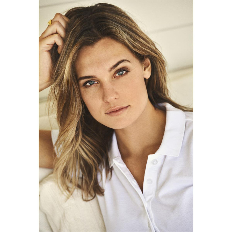 A woman with long brown hair is wearing a white polo shirt, posing against a light background.