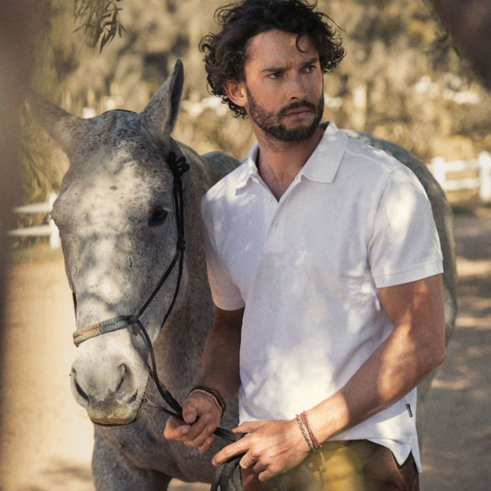A man wearing a white cotton polo shirt stands beside a grey horse, set in a natural outdoor environment.