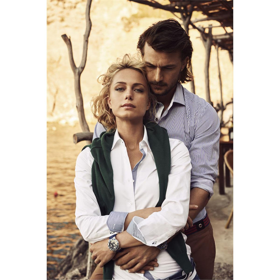 A model wears a white button-up blouse, layered with a green sweater, against a scenic outdoor backdrop.