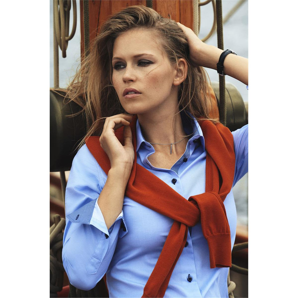 A woman in a blue button-up blouse with an orange sweater tied around her shoulders, set against a nautical backdrop.
