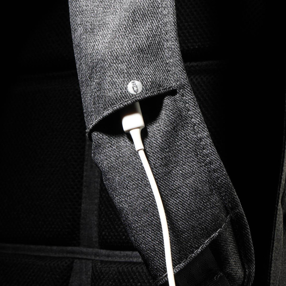 A close-up of a black backpack strap featuring a white charging cable tucked inside. The strap has a logo.