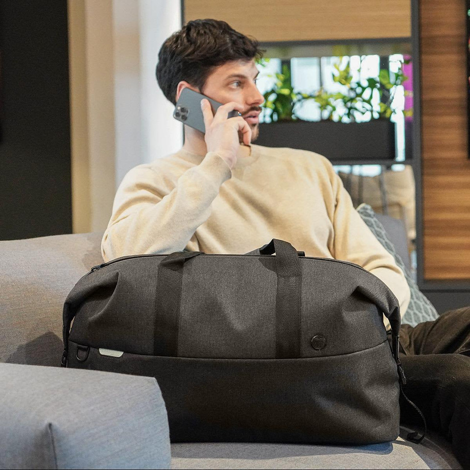 A grey duffle bag sits beside a person on a couch, who is talking on the phone. The bag features a logo.