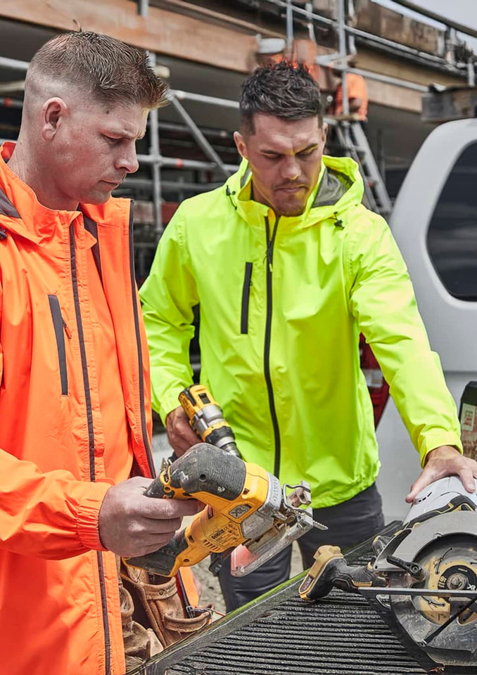 Two men in bright orange and neon yellow rain jackets work on a construction site, using power tools. Both jackets have logos.