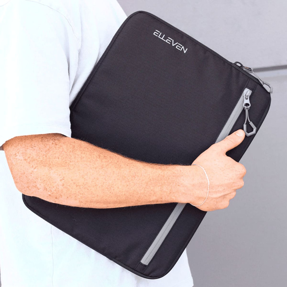A laptop sleeve in black with a grey zipper, featuring a logo, held under someone's arm.